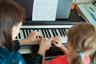 A person is teaching a child how to play the piano, with sheet music placed on top of the piano. The hands of the adult are gently guiding the child's fingers over the keys.