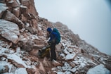 A person dressed in hiking gear stands on a rocky mountain path that is dusted with snow. The hiker is wearing a blue jacket and carrying a backpack, and appears to be adjusting equipment or unpacking. The mountainside is steep and rugged, with a foggy sky above.