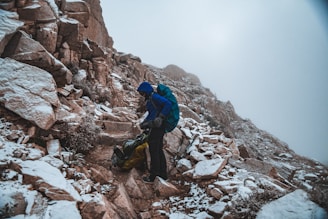 A person dressed in hiking gear stands on a rocky mountain path that is dusted with snow. The hiker is wearing a blue jacket and carrying a backpack, and appears to be adjusting equipment or unpacking. The mountainside is steep and rugged, with a foggy sky above.