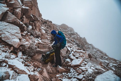 A person dressed in hiking gear stands on a rocky mountain path that is dusted with snow. The hiker is wearing a blue jacket and carrying a backpack, and appears to be adjusting equipment or unpacking. The mountainside is steep and rugged, with a foggy sky above.