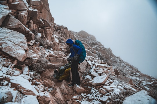 A person dressed in hiking gear stands on a rocky mountain path that is dusted with snow. The hiker is wearing a blue jacket and carrying a backpack, and appears to be adjusting equipment or unpacking. The mountainside is steep and rugged, with a foggy sky above.