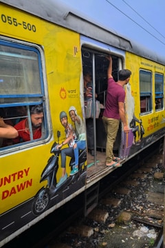A train carriage with a bright yellow advertisement on its exterior, depicting a couple riding a scooter. Several passengers are inside, holding onto the metal handles, with one man standing and partially leaning out of the open door. The train is stationary on a track with gravel underneath.