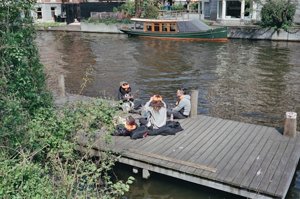 A group of people is sitting on a wooden dock by a canal, with greenery surrounding the area. They appear to be engaged in conversation, wearing casual clothing and some adorned with orange accessories. A small wooden boat is moored in the canal, and buildings are visible in the background.