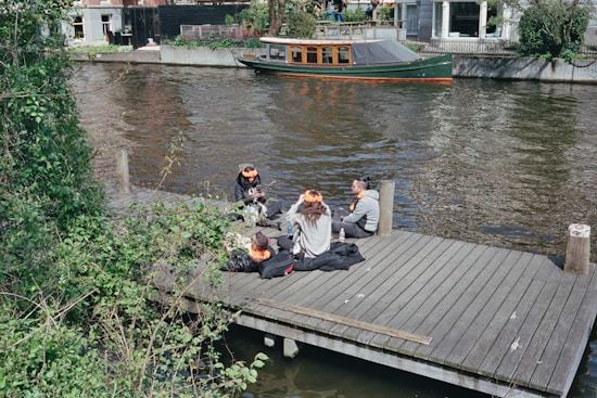 A group of people is sitting on a wooden dock by a canal, with greenery surrounding the area. They appear to be engaged in conversation, wearing casual clothing and some adorned with orange accessories. A small wooden boat is moored in the canal, and buildings are visible in the background.