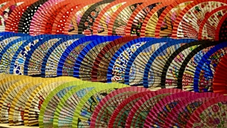 A vibrant display of traditional hand fans arranged in several rows. The fans are decorated with intricate patterns and diverse colors, such as polka dots and floral designs. Each row features a distinct color palette, including shades of red, blue, green, and yellow.