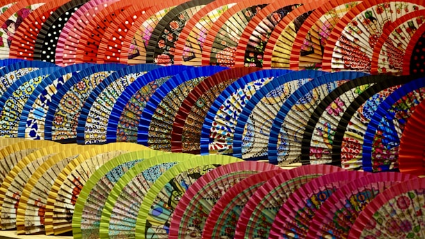 A vibrant display of traditional hand fans arranged in several rows. The fans are decorated with intricate patterns and diverse colors, such as polka dots and floral designs. Each row features a distinct color palette, including shades of red, blue, green, and yellow.
