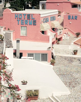 A vibrant, pastel-toned hotel complex with the names 'Hotel Terme' and 'Casa Rosa' displayed on the buildings. The architecture features pink walls and terraces adorned with potted plants and flowers. The courtyard has a mosaic-patterned path, and there is a bar area visible. The scene is bathed in bright sunlight, casting sharp shadows.