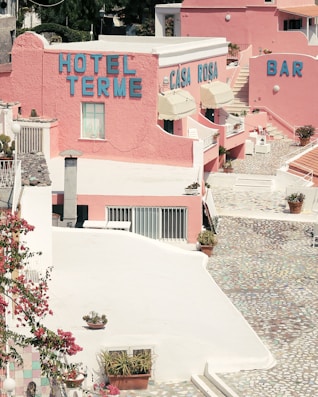 A vibrant, pastel-toned hotel complex with the names 'Hotel Terme' and 'Casa Rosa' displayed on the buildings. The architecture features pink walls and terraces adorned with potted plants and flowers. The courtyard has a mosaic-patterned path, and there is a bar area visible. The scene is bathed in bright sunlight, casting sharp shadows.