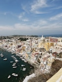 An aerial view of a colorful, coastal Italian town with numerous pastel-colored buildings clustered along the curving shoreline. Small boats are docked in the harbor, and the deep blue sea stretches beyond. The sky is mostly clear with a few wispy clouds.