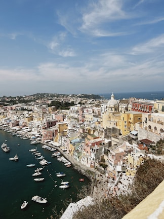 An aerial view of a colorful, coastal Italian town with numerous pastel-colored buildings clustered along the curving shoreline. Small boats are docked in the harbor, and the deep blue sea stretches beyond. The sky is mostly clear with a few wispy clouds.