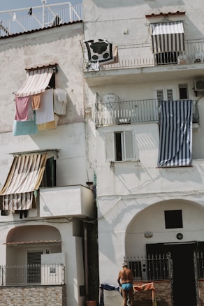 A multi-story residential building with balconies, where various colorful clothes and towels are hung out to dry. A person in swimwear stands at the lower level near the entrance. The atmosphere appears relaxed and everyday.