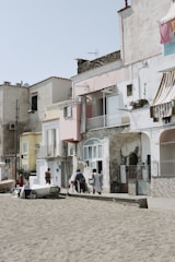 A seaside scene with a row of colorful, weathered houses lining the beachfront. Several people are gathered near the entrance of one house, engaged in conversation. There are small boats positioned on the sand nearby, and clothes hang to dry from balconies.