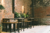 A rustic outdoor seating area features multiple wooden bar tables and stools against a backdrop of exposed brick walls. Green potted plants add a touch of nature to the otherwise industrial setting, with soft lighting providing a cozy and intimate atmosphere.