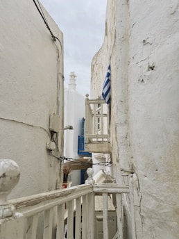 Narrow alleyway between whitewashed buildings with a small balcony displaying a blue and white striped flag. The architectural style is simple and rustic, characterized by chipped paint and exposed brick. Electrical wires are visible, adding to the urban charm.