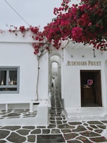 A narrow, cobblestone path lined with white-washed buildings and vibrant pink bougainvillea vines. The architecture features an entrance to a luxury store named Audemars Piguet with minimalistic and clean design elements.