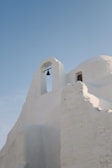 An architectural structure with smooth, white walls and a distinct arch holding a small bell. The building exhibits traditional Cycladic style, characterized by organic shapes and soft lines. Above, a clear blue sky enhances its serene and minimalistic beauty.