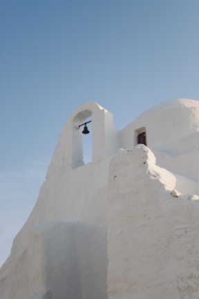 An architectural structure with smooth, white walls and a distinct arch holding a small bell. The building exhibits traditional Cycladic style, characterized by organic shapes and soft lines. Above, a clear blue sky enhances its serene and minimalistic beauty.