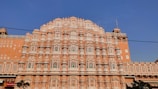 An ornate, multi-level palace facade with a series of intricately carved pink sandstone windows, traditional in Rajasthani architecture. Symmetrical design with hundreds of small windows decorated with elaborate latticework, against a clear blue sky.