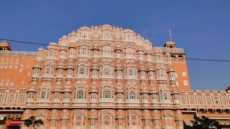 An ornate, multi-level palace facade with a series of intricately carved pink sandstone windows, traditional in Rajasthani architecture. Symmetrical design with hundreds of small windows decorated with elaborate latticework, against a clear blue sky.