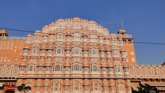 An ornate, multi-level palace facade with a series of intricately carved pink sandstone windows, traditional in Rajasthani architecture. Symmetrical design with hundreds of small windows decorated with elaborate latticework, against a clear blue sky.