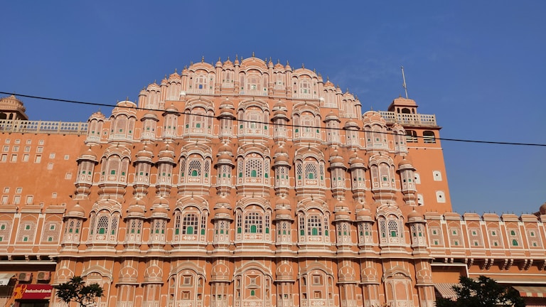 An ornate, multi-level palace facade with a series of intricately carved pink sandstone windows, traditional in Rajasthani architecture. Symmetrical design with hundreds of small windows decorated with elaborate latticework, against a clear blue sky.