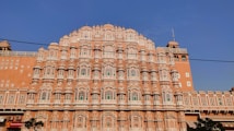 An ornate, multi-level palace facade with a series of intricately carved pink sandstone windows, traditional in Rajasthani architecture. Symmetrical design with hundreds of small windows decorated with elaborate latticework, against a clear blue sky.