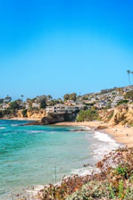 Coastal scene featuring a pristine sandy beach with turquoise ocean waters gently lapping at the shore. In the background, several houses and buildings are nestled on cliffs, surrounded by lush greenery. A clear blue sky stretches above, adding to the tranquil atmosphere. Sparse beachgoers can be seen enjoying the sunlight and the ocean view.