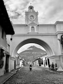 A picturesque colonial archway with a clock tower spans a cobblestone street. In the background, a volcano is visible under a cloudy sky. People are walking and a motorcyclist is riding down the street, lined with traditional buildings.