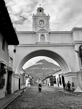 A picturesque colonial archway with a clock tower spans a cobblestone street. In the background, a volcano is visible under a cloudy sky. People are walking and a motorcyclist is riding down the street, lined with traditional buildings.