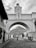 A picturesque colonial archway with a clock tower spans a cobblestone street. In the background, a volcano is visible under a cloudy sky. People are walking and a motorcyclist is riding down the street, lined with traditional buildings.