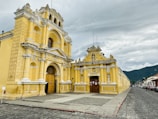 A historic yellow colonial-style building with intricate architectural details stands prominently beside a cobblestone street. The façade features ornate white trim, arched doorways, and sculptures embedded in the structure. A cloudy sky looms above, with mountains visible in the background.