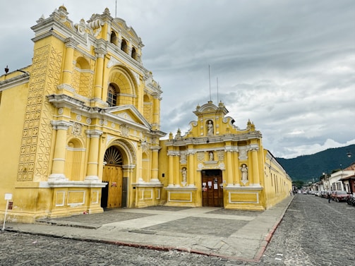 A historic yellow colonial-style building with intricate architectural details stands prominently beside a cobblestone street. The façade features ornate white trim, arched doorways, and sculptures embedded in the structure. A cloudy sky looms above, with mountains visible in the background.