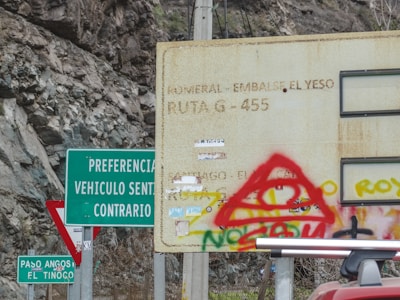 Several road signs are located in a mountainous area with rocky terrain. A large rusted sign is partially obscured by red graffiti, while other signs include green and white directional indicators. Some signs are clearer than others, and there