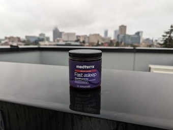 A jar labeled 'medterra Fast Asleep' sits on a wet surface with a blurred urban skyline in the background. The jar is dark with purple labeling, indicating it contains 2mg CBN gummies. The scene is overcast, suggesting a rainy day.