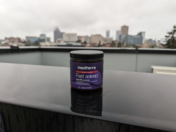 A jar labeled 'medterra Fast Asleep' sits on a wet surface with a blurred urban skyline in the background. The jar is dark with purple labeling, indicating it contains 2mg CBN gummies. The scene is overcast, suggesting a rainy day.