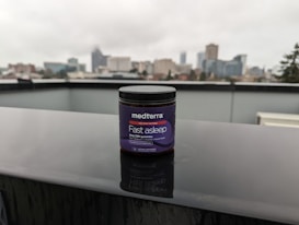 A jar labeled 'medterra Fast Asleep' sits on a wet surface with a blurred urban skyline in the background. The jar is dark with purple labeling, indicating it contains 2mg CBN gummies. The scene is overcast, suggesting a rainy day.