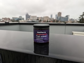 A jar labeled 'medterra Fast Asleep' sits on a wet surface with a blurred urban skyline in the background. The jar is dark with purple labeling, indicating it contains 2mg CBN gummies. The scene is overcast, suggesting a rainy day.