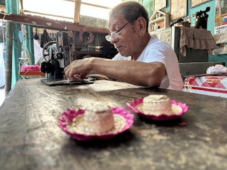 An elderly man wearing glasses is focused on sewing with a vintage sewing machine in a rustic and colorful room. The table in the foreground holds small decorative hats with pink trims. The background features various household items and wall hangings that add a sense of lived-in warmth.
