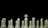 A row of white chess pieces is lined up against a black background. The pieces include a king at the center, marked by a cross on top, surrounded by other pieces like rooks, knights, bishops, and pawns. The arrangement and lighting highlight the smooth texture and shapes of the chess pieces.