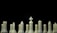 A row of white chess pieces is lined up against a black background. The pieces include a king at the center, marked by a cross on top, surrounded by other pieces like rooks, knights, bishops, and pawns. The arrangement and lighting highlight the smooth texture and shapes of the chess pieces.