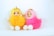 Two cute plush dolls sitting side by side, each with a detailed face and dressed in a fluffy outfit. The left doll is adorned in yellow with white limbs and a golden bow on its head. The right doll wears a pink outfit with similar white limbs and a golden bow on its head. Both dolls have expressive painted faces and are placed against a plain white background.