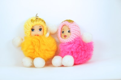 Two cute plush dolls sitting side by side, each with a detailed face and dressed in a fluffy outfit. The left doll is adorned in yellow with white limbs and a golden bow on its head. The right doll wears a pink outfit with similar white limbs and a golden bow on its head. Both dolls have expressive painted faces and are placed against a plain white background.