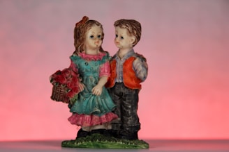 Two figurines of a boy and a girl standing closely together. The girl is wearing a teal dress with pink accents and holding a basket of red flowers. The boy is dressed in a gray shirt with a bright orange vest and dark pants. The background features a gradient of soft pink and light purple tones.