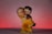 A small figurine of a couple in a loving embrace, with the man holding the woman as they both gaze lovingly at each other. The woman wears a yellow dress and has a purple bow in her hair, while the man is dressed in darker clothing. A single red rose is visible in the woman's hand. The background features a smooth gradient of soft pink to dark red.