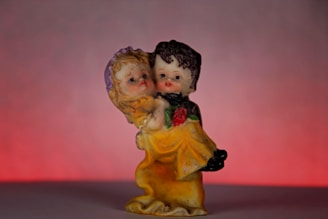A small figurine of a couple in a loving embrace, with the man holding the woman as they both gaze lovingly at each other. The woman wears a yellow dress and has a purple bow in her hair, while the man is dressed in darker clothing. A single red rose is visible in the woman's hand. The background features a smooth gradient of soft pink to dark red.