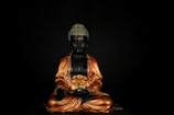 A statue of a sitting Buddha with a serene expression, adorned in ornate golden robes against a dark background. The Buddha's hands are placed in a meditative gesture with intricate detailing on the robe.