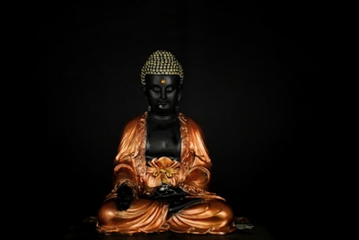 A statue of a sitting Buddha with a serene expression, adorned in ornate golden robes against a dark background. The Buddha's hands are placed in a meditative gesture with intricate detailing on the robe.