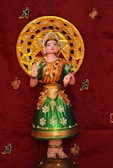 A colorful traditional Indian doll is depicted wearing an ornate costume, adorned with shimmering jewels and a vivid green skirt embellished with floral patterns. The doll stands against a rich, deep red backdrop with golden and green decorative elements. It features an elaborate golden halo behind its head, contributing to a sense of divinity or cultural significance.