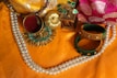 A collection of traditional jewelry and ornaments displayed on a vibrant orange fabric. The arrangement features a double-stranded pearl necklace, two colorful bangles in green and gold, a circular brooch with turquoise embellishments, a small round container holding red colored powder, and a decorative box. A pink and gold fabric is partially visible in the background, adding richness to the composition.