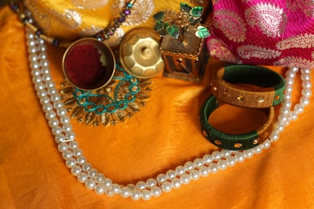 A collection of traditional jewelry and ornaments displayed on a vibrant orange fabric. The arrangement features a double-stranded pearl necklace, two colorful bangles in green and gold, a circular brooch with turquoise embellishments, a small round container holding red colored powder, and a decorative box. A pink and gold fabric is partially visible in the background, adding richness to the composition.